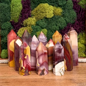Puede incluir: Una colección de once torres de piedra pulida con puntas afiladas, que presentan una variedad de colores que incluyen rojo intenso, amarillo mostaza, crema y morado, dispuestas en grupo sobre una superficie de madera con un fondo de musgo verde y morado.