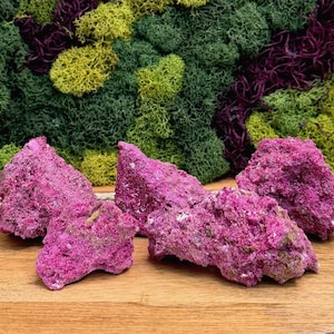 May include: Four jagged, vibrant magenta mineral specimens with a crystalline, rough texture are arranged on a smooth, light-toned wooden surface. The rocks feature deep pink hues and irregular, sharp edges, contrasting with the natural wood grain.