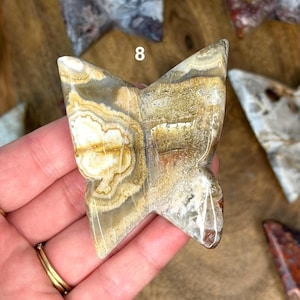 May include: A butterfly-shaped stone carving with a natural, earthy color palette of beige, brown, and white. The stone has a polished surface and visible veining, with the number 8 visible on the top. The carving is held in a hand.