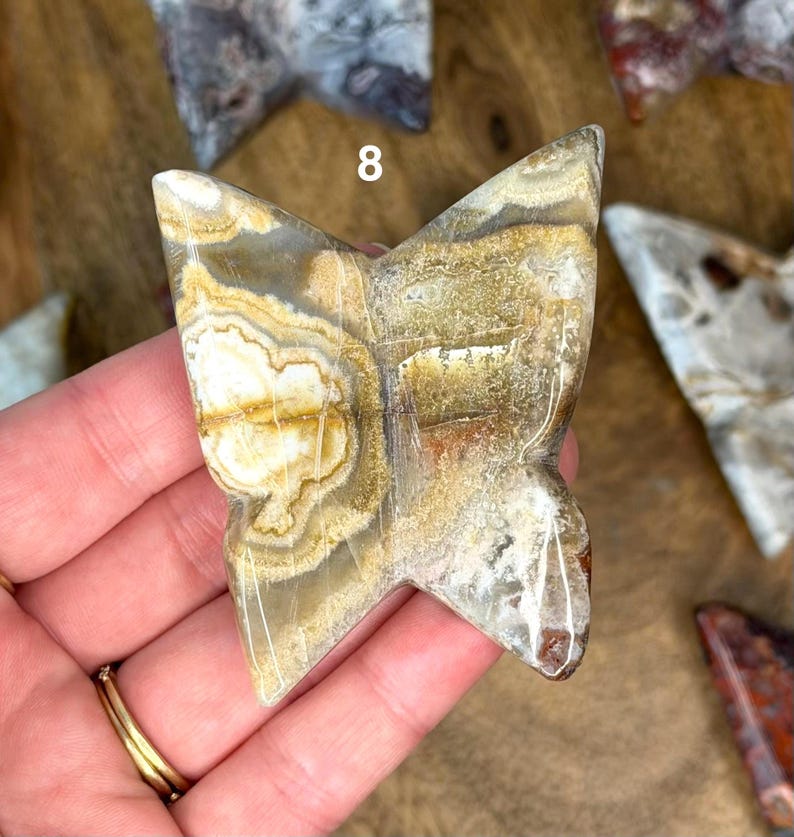 May include: A butterfly-shaped stone carving with a natural, earthy color palette of beige, brown, and white. The stone has a polished surface and visible veining, with the number 8 visible on the top. The carving is held in a hand.