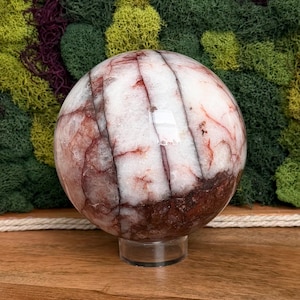 May include: A large, polished sphere with a mix of white, red, and brown tones. The orb is supported by a clear, circular stand. The sphere's surface has a marbled appearance, with veins of red and brown running through the white base. The background features green moss.