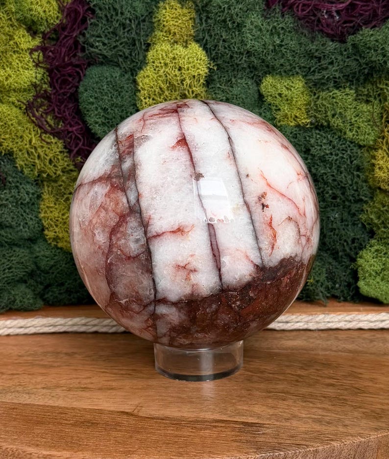 May include: A large, polished sphere with a mix of white, red, and brown tones. The orb is supported by a clear, circular stand. The sphere's surface has a marbled appearance, with veins of red and brown running through the white base. The background features green moss.