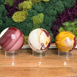 Puede incluir: Tres esferas de piedra pulida sobre soportes circulares transparentes. La esfera 6 es de color granate oscuro con bandas blancas. La esfera 7 es crema con remolinos granates y dorados. La esfera 8 es amarillo dorado con vetas rojas y marrones.