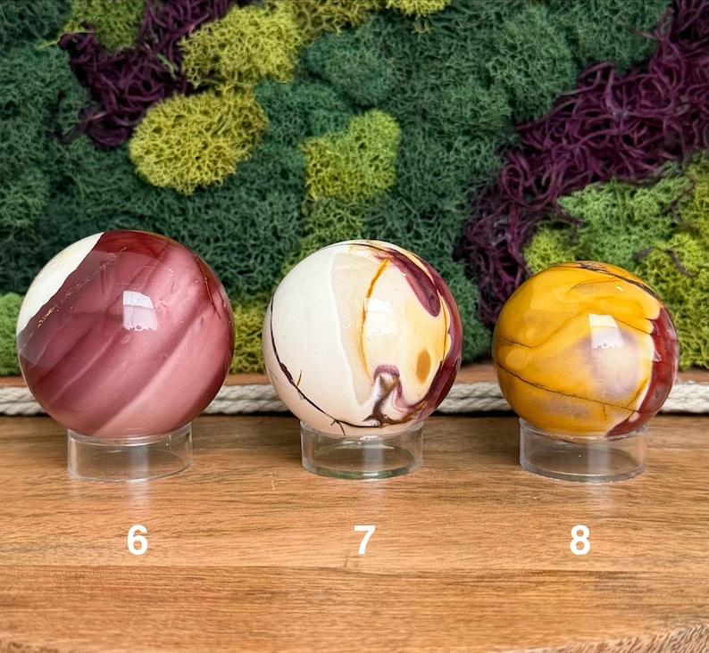 Puede incluir: Tres esferas de piedra pulida sobre soportes circulares transparentes. La esfera 6 es de color granate oscuro con bandas blancas. La esfera 7 es crema con remolinos granates y dorados. La esfera 8 es amarillo dorado con vetas rojas y marrones.