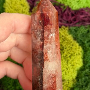 May include: A clear, reddish-brown crystal tower with a pointed top. The crystal has internal veining and inclusions, and the number "10" is visible on the top. The background features green and purple moss.