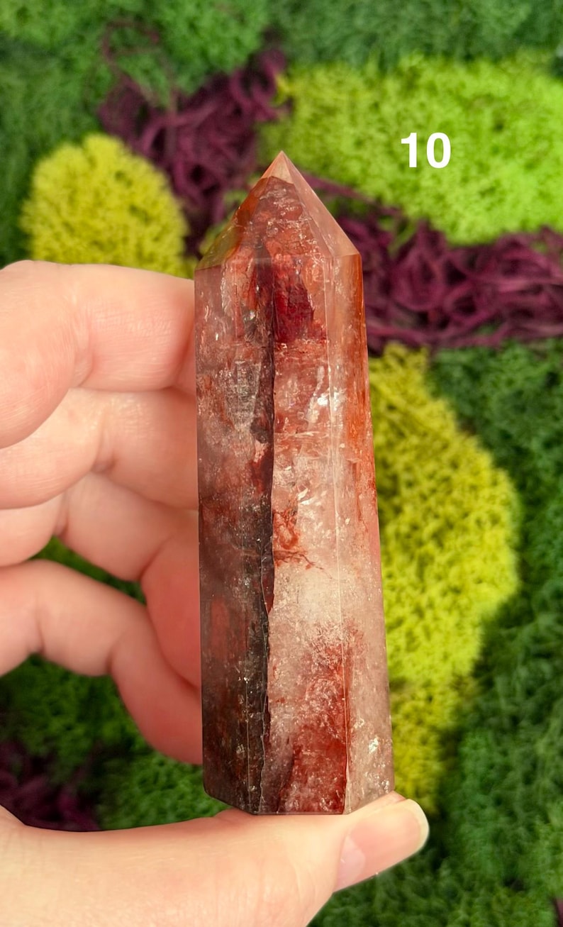 May include: A clear, reddish-brown crystal tower with a pointed top. The crystal has internal veining and inclusions, and the number "10" is visible on the top. The background features green and purple moss.
