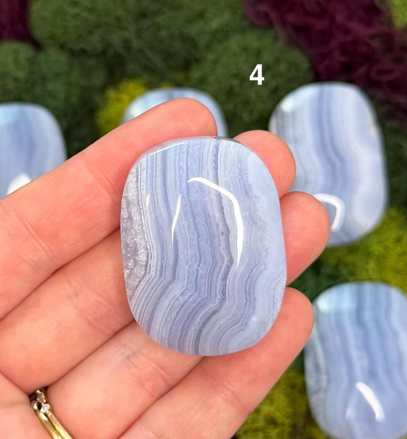 May include: A collection of polished blue lace agate stones. The stones have a smooth, oval shape with light blue and white banding patterns. The number "4" is visible in the background. The stones are displayed on a bed of green moss.