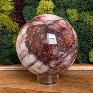 May include: A large, polished crystal sphere with a mix of white and deep red veining. The sphere is displayed on a clear, round stand, set against a backdrop of green and purple moss. The crystal ball has a reflective surface.