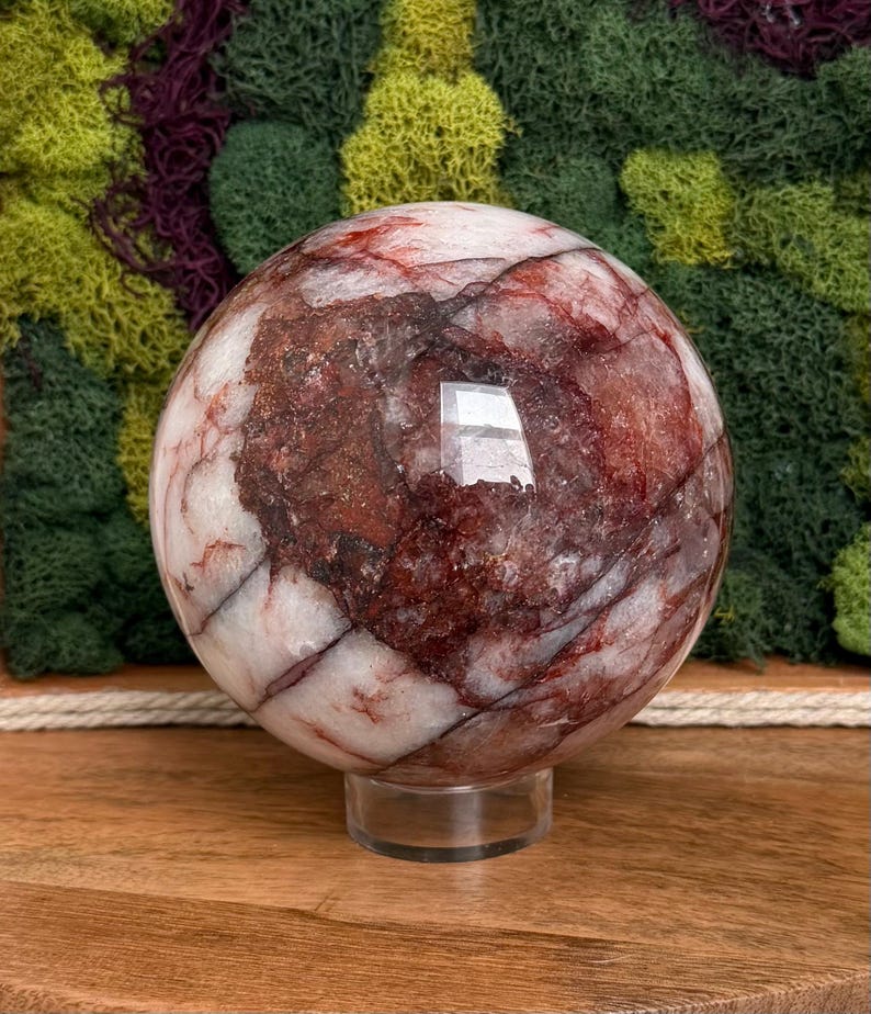 May include: A large, polished crystal sphere with a mix of white and deep red veining. The sphere is displayed on a clear, round stand, set against a backdrop of green and purple moss. The crystal ball has a reflective surface.