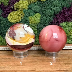 Puede incluir: Dos esferas de piedra pulida sobre soportes circulares transparentes. La esfera 1 presenta un patr&oacute;n multicolor de bandas blancas, amarillas y rojo oscuro con inclusiones. La esfera 2 es de un color rosa viejo uniforme, liso y s&oacute;lido.
