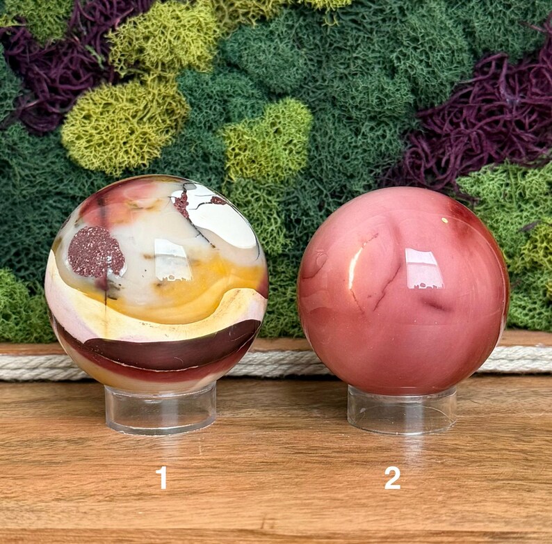 Puede incluir: Dos esferas de piedra pulida sobre soportes circulares transparentes. La esfera 1 presenta un patr&oacute;n multicolor de bandas blancas, amarillas y rojo oscuro con inclusiones. La esfera 2 es de un color rosa viejo uniforme, liso y s&oacute;lido.