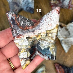 May include: A carved stone butterfly sculpture with a mix of colors including white, black, brown, and blue. The sculpture is held in a hand, and the number "10" is visible on the sculpture. The butterfly has a detailed, layered pattern.