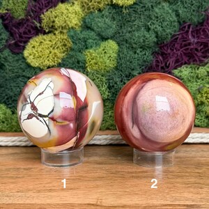 Puede incluir: Dos piedras esf&eacute;ricas pulidas sobre soportes circulares transparentes. La esfera izquierda presenta patrones blancos, rojo oscuro y amarillos con vetas oscuras. La esfera derecha muestra bandas circulares conc&eacute;ntricas en tonos rojo, rosa y beige.