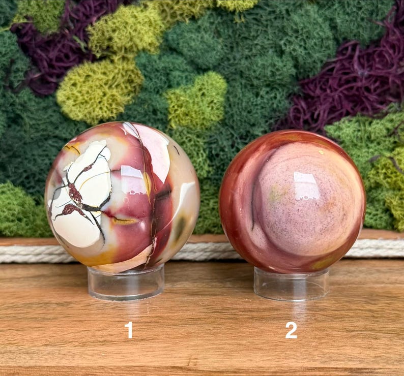 Puede incluir: Dos piedras esf&eacute;ricas pulidas sobre soportes circulares transparentes. La esfera izquierda presenta patrones blancos, rojo oscuro y amarillos con vetas oscuras. La esfera derecha muestra bandas circulares conc&eacute;ntricas en tonos rojo, rosa y beige.