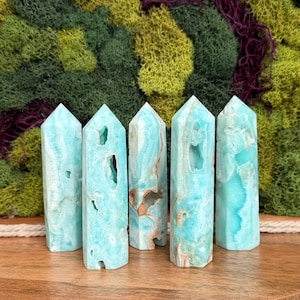 May include: Five blue-green crystal towers with varying patterns and internal cavities. The crystals are standing upright on a wooden surface, with a backdrop of green and purple moss.