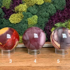 Puede incluir: Tres piedras esf&eacute;ricas pulidas sobre soportes circulares transparentes. La primera esfera es roja y amarilla, la segunda es de color malva s&oacute;lido con vetas oscuras y la tercera es una mezcla de morado oscuro, crema y rojo. Etiquetadas 3, 4 y 5.
