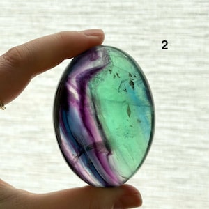 Puede incluir: Una piedra de fluorita pulida, de forma ovalada. La piedra presenta una mezcla de colores, incluyendo verde, morado y azul, con un aspecto transl&uacute;cido. El n&uacute;mero "2" es visible en la piedra.