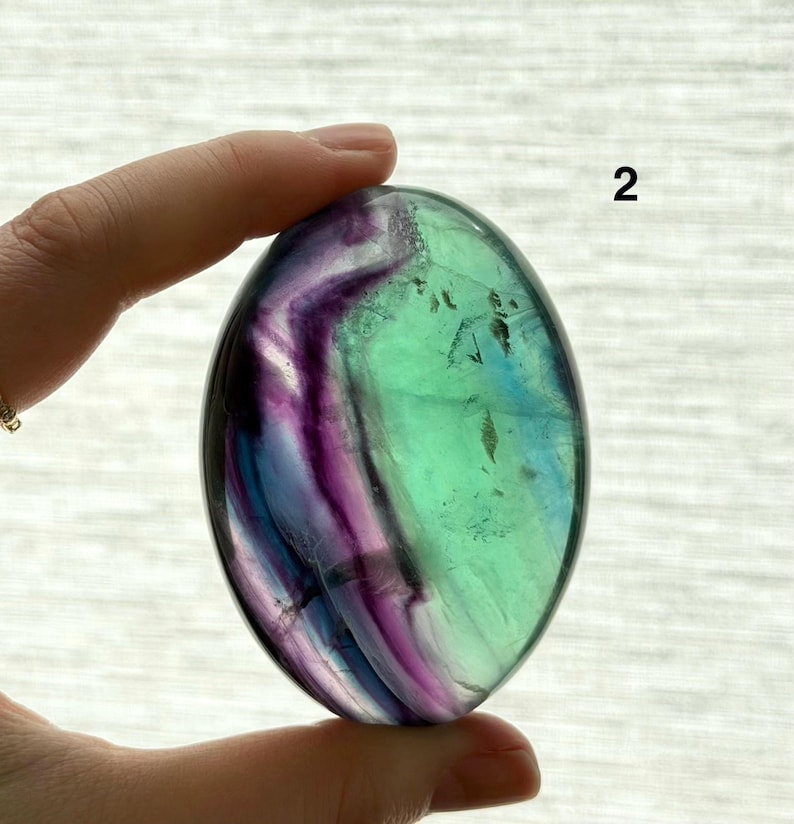 Puede incluir: Una piedra de fluorita pulida, de forma ovalada. La piedra presenta una mezcla de colores, incluyendo verde, morado y azul, con un aspecto transl&uacute;cido. El n&uacute;mero "2" es visible en la piedra.