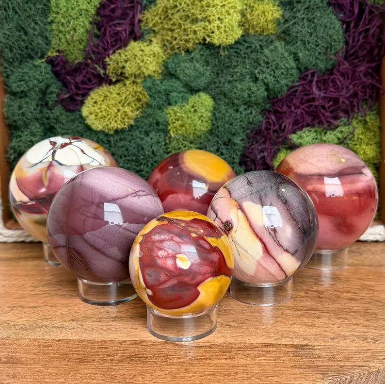 Puede incluir: Una colecci&oacute;n de cinco piedras esf&eacute;ricas pulidas con patrones de remolinos en tonos p&uacute;rpura intenso, amarillo mostaza, crema y granate. Cada orbe mineral liso y redondeado se exhibe sobre un peque&ntilde;o soporte circular transparente.