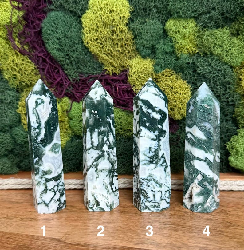 May include: Four polished moss agate crystal towers. Each tower is a different size and shape, with a white and green pattern. The crystals are numbered 1 through 4. The background is a mix of green, yellow, and purple moss.