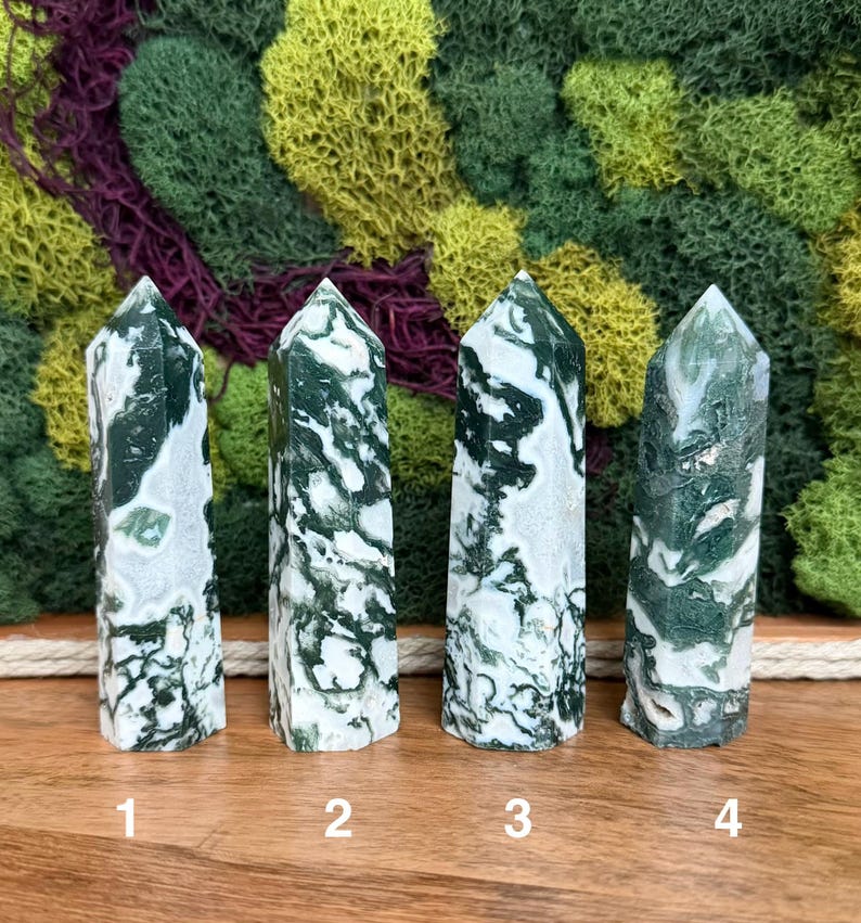 May include: Four green and white moss agate crystal points are displayed on a wooden surface. The crystals are numbered 1 through 4. The background features a mix of green, yellow, and purple moss.