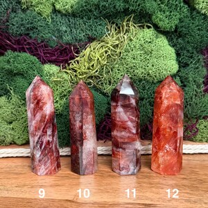 May include: Four red quartz crystal points with varying shades of red and white inclusions. Each crystal is a different size and shape, with a pointed top. The crystals are numbered 9, 10, 11, and 12. The background features green and purple moss.