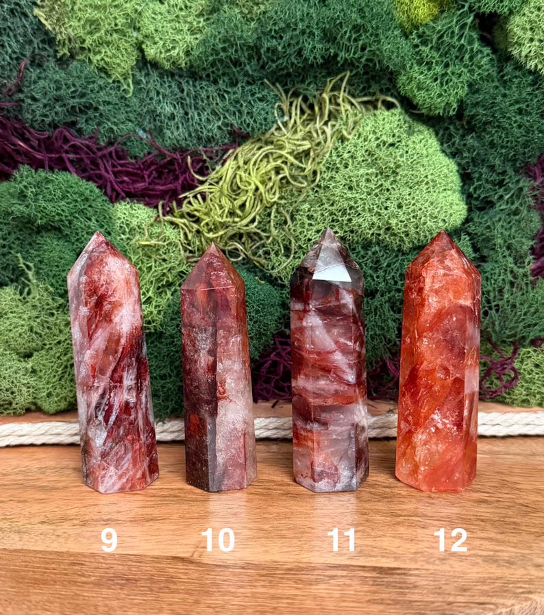 May include: Four red quartz crystal points with varying shades of red and white inclusions. Each crystal is a different size and shape, with a pointed top. The crystals are numbered 9, 10, 11, and 12. The background features green and purple moss.