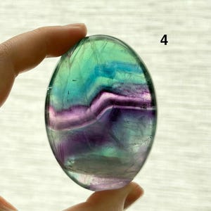 Puede incluir: Una piedra de fluorita ovalada pulida. La piedra presenta una mezcla de colores, incluyendo verde, azul y morado, con una calidad transl&uacute;cida. La piedra est&aacute; sujeta por una mano, y el n&uacute;mero "4" es visible en el fondo.