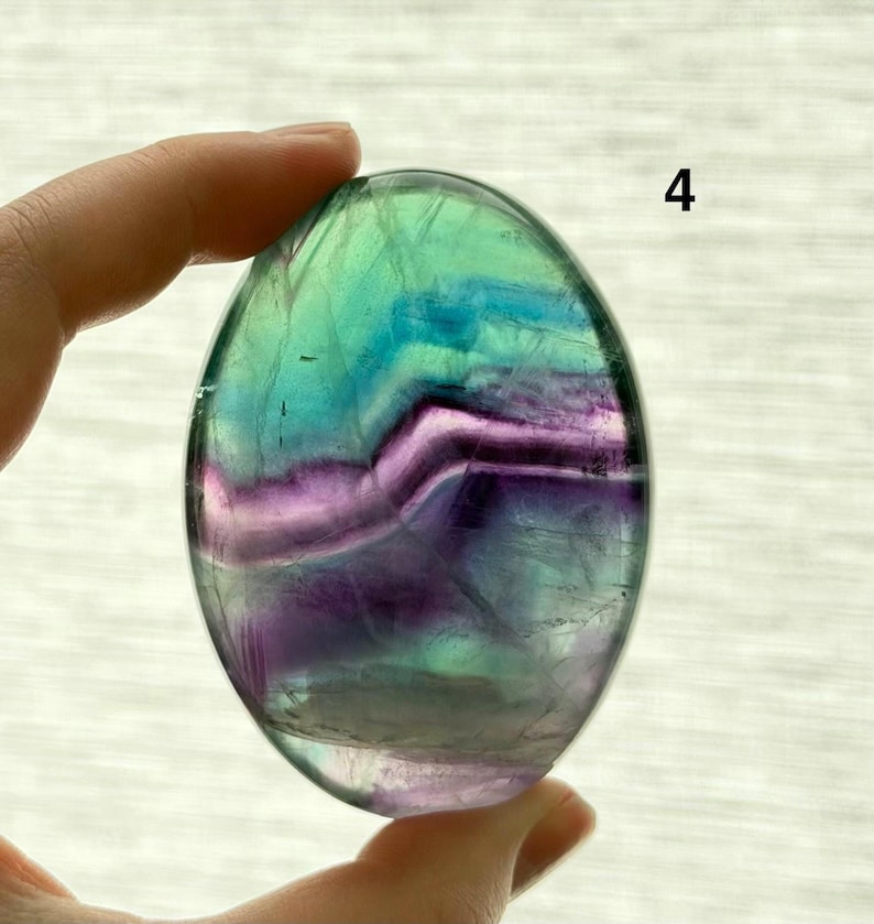 Puede incluir: Una piedra de fluorita ovalada pulida. La piedra presenta una mezcla de colores, incluyendo verde, azul y morado, con una calidad transl&uacute;cida. La piedra est&aacute; sujeta por una mano, y el n&uacute;mero "4" es visible en el fondo.