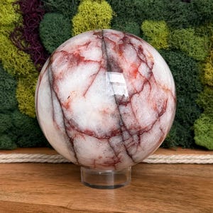 May include: A polished, spherical gemstone with a white base and striking red veining. The orb is displayed on a clear, circular stand, showcasing its natural patterns. The background features a textured display of green and purple moss.
