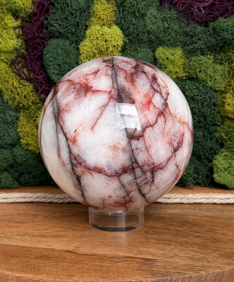 May include: A polished, spherical gemstone with a white base and striking red veining. The orb is displayed on a clear, circular stand, showcasing its natural patterns. The background features a textured display of green and purple moss.