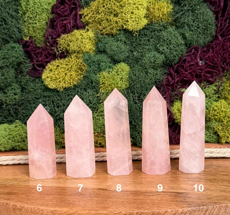 May include: Five light pink rose quartz crystal points arranged in a row on a wooden surface. Each point is numbered 6 through 10 at the base, showing a faceted top and a smooth, translucent body with natural internal inclusions and variations.