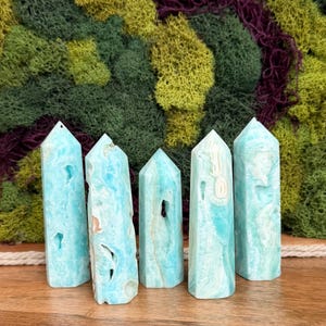 May include: Five blue-green crystal towers with varying patterns and textures. The crystals are standing upright on a wooden surface, with a backdrop of green and purple moss. Each crystal has a pointed top and a rectangular base.