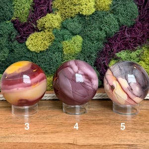 Puede incluir: Tres piedras esf&eacute;ricas pulidas sobre soportes circulares transparentes. La esfera 3 presenta bandas amarillas, rojas y moradas. La esfera 4 es de un morado intenso con vetas sutiles. La esfera 5 muestra una mezcla de remolinos morados, crema y gris oscuro.