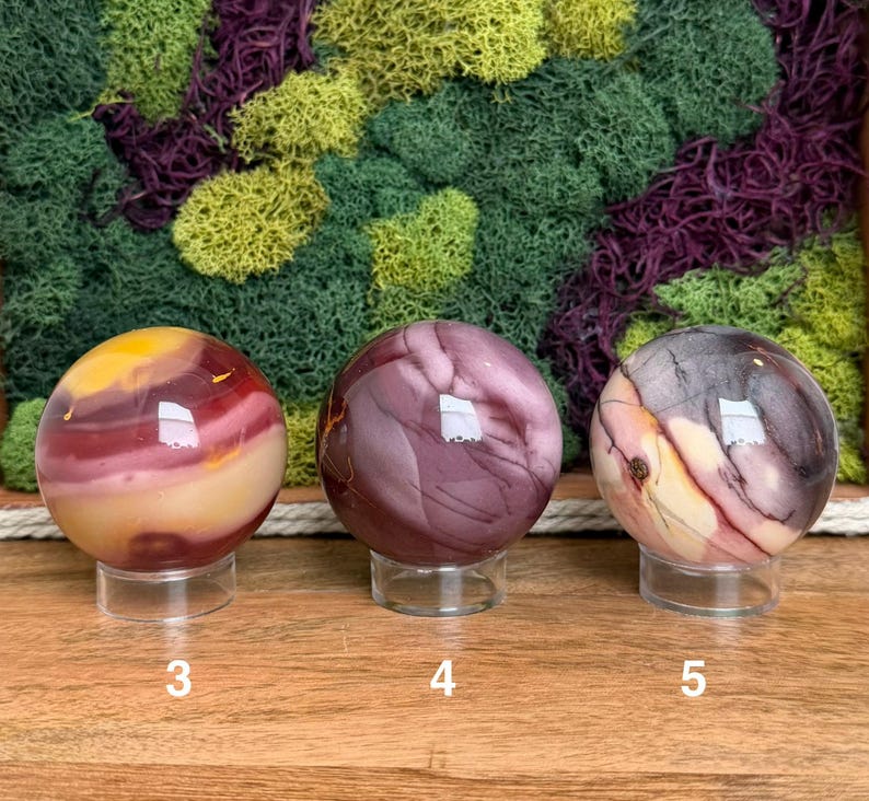 Puede incluir: Tres piedras esf&eacute;ricas pulidas sobre soportes circulares transparentes. La esfera 3 presenta bandas amarillas, rojas y moradas. La esfera 4 es de un morado intenso con vetas sutiles. La esfera 5 muestra una mezcla de remolinos morados, crema y gris oscuro.