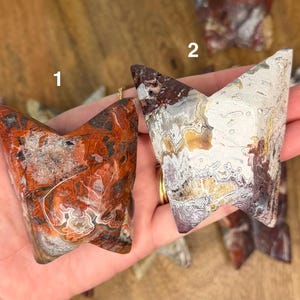 May include: Two polished, V-shaped agate stones. One is primarily red and brown with white and gray inclusions. The other is white, gray, and brown with a marbled appearance. Both are held in a hand.