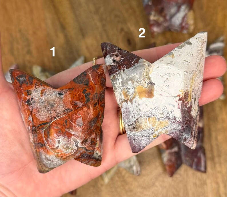 May include: Two polished, V-shaped agate stones. One is primarily red and brown with white and gray inclusions. The other is white, gray, and brown with a marbled appearance. Both are held in a hand.