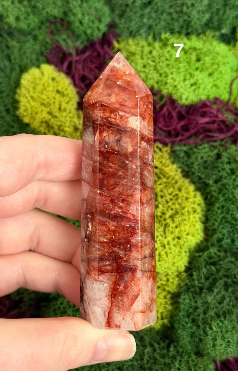 May include: A red and clear quartz crystal point with a polished, faceted surface. The crystal features reddish-brown inclusions and a pointed top. The background is a mix of green and purple moss.