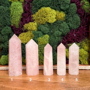 May include: Five polished rose quartz crystal towers arranged in a row on a wooden surface. Each tower features a hexagonal point and a pale pink, translucent appearance. The crystals are numbered one through five in white text at the base.