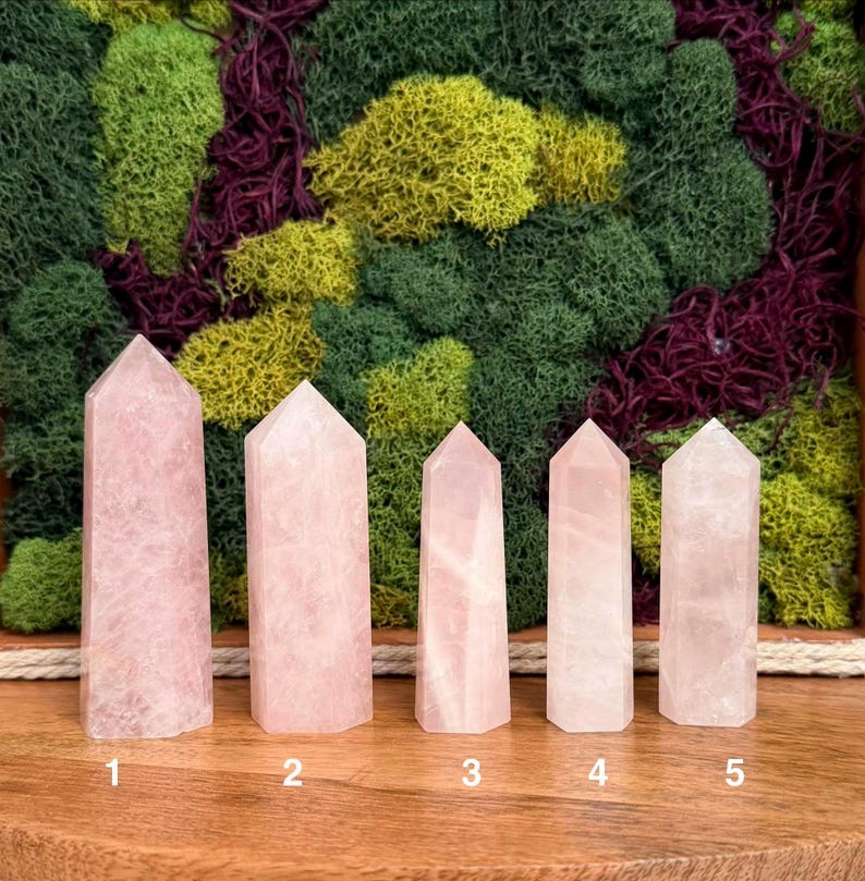 May include: Five polished rose quartz crystal towers arranged in a row on a wooden surface. Each tower features a hexagonal point and a pale pink, translucent appearance. The crystals are numbered one through five in white text at the base.
