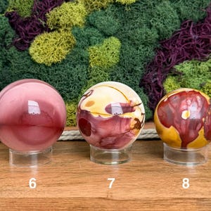 Puede incluir: Tres piedras esf&eacute;ricas pulidas sobre soportes circulares transparentes, etiquetadas 6, 7 y 8. La primera es de color rosa empolvado s&oacute;lido, la segunda presenta remolinos crema y granate, y la tercera muestra una mezcla de rojo, amarillo y marr&oacute;n.
