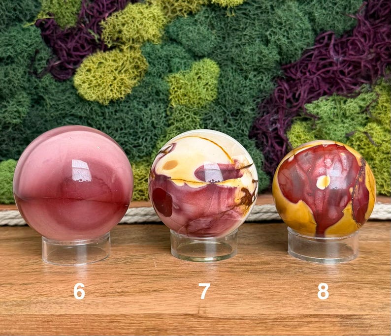 Puede incluir: Tres piedras esf&eacute;ricas pulidas sobre soportes circulares transparentes, etiquetadas 6, 7 y 8. La primera es de color rosa empolvado s&oacute;lido, la segunda presenta remolinos crema y granate, y la tercera muestra una mezcla de rojo, amarillo y marr&oacute;n.
