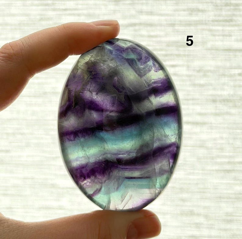 Puede incluir: Una piedra de fluorita ovalada con una superficie pulida. La piedra presenta bandas de colores p&uacute;rpura, turquesa y claros. El n&uacute;mero "5" es visible en la esquina superior derecha. La piedra se sostiene entre dos dedos.
