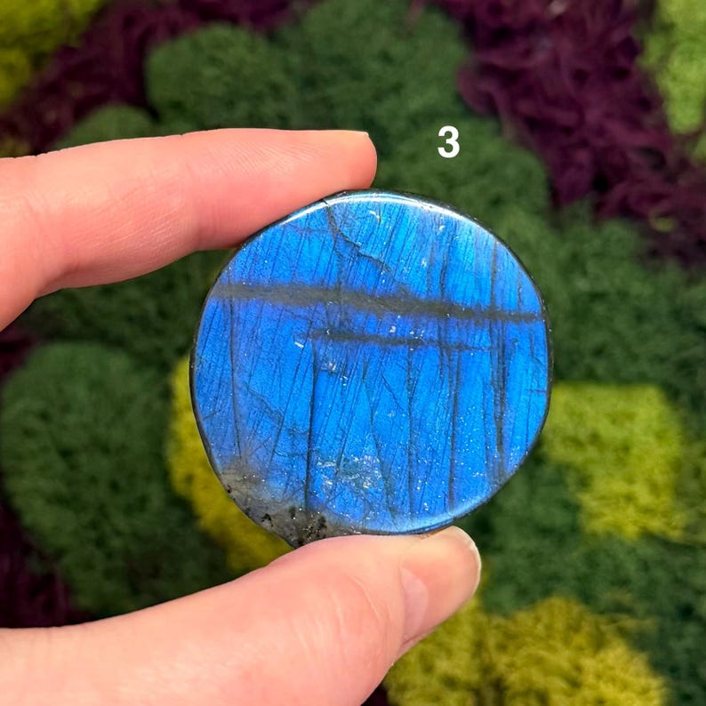 May include: A round, blue labradorite stone with dark gray veining. The stone is held in a hand and the number "3" is visible in the background. The stone has a polished surface and displays a play of color.