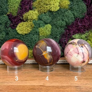 Puede incluir: Tres esferas de piedra pulida sobre soportes circulares transparentes. La esfera 3 es roja y amarilla, la 4 es marr&oacute;n oscuro con vetas doradas y la 5 es una mezcla de rosa, crema y marr&oacute;n oscuro. Cada esfera tiene un n&uacute;mero blanco.