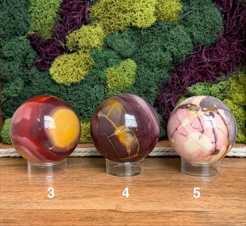 Puede incluir: Tres esferas de piedra pulida sobre soportes circulares transparentes. La esfera 3 es roja y amarilla, la 4 es marr&oacute;n oscuro con vetas doradas y la 5 es una mezcla de rosa, crema y marr&oacute;n oscuro. Cada esfera tiene un n&uacute;mero blanco.
