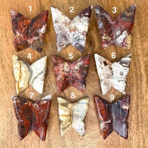 May include: Nine carved stone butterfly figurines in various colors, including red, brown, white, and beige. Each butterfly has unique patterns and textures, displayed on a wooden surface. The butterflies are numbered 1 through 9.
