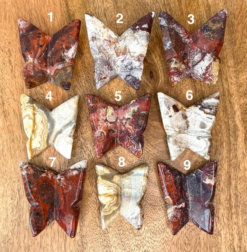 May include: Nine carved stone butterfly figurines in various colors, including red, brown, white, and beige. Each butterfly has unique patterns and textures, displayed on a wooden surface. The butterflies are numbered 1 through 9.