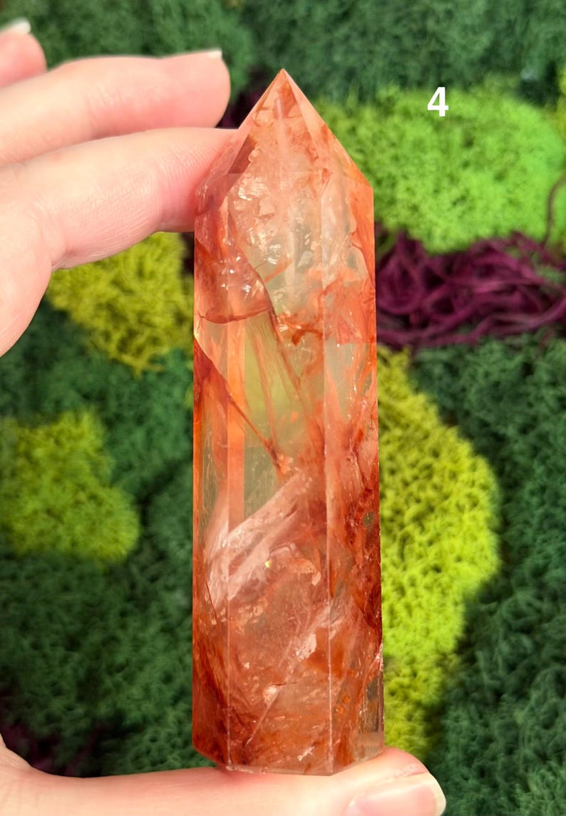 May include: A clear quartz crystal point with reddish-orange inclusions. The crystal is held against a backdrop of green and purple moss. The number "4" is visible in the background.