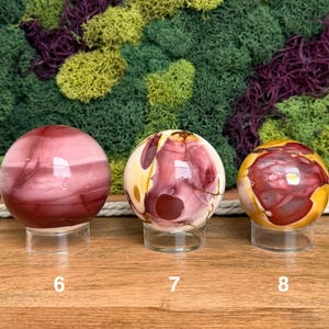 Puede incluir: Tres piedras esf&eacute;ricas pulidas sobre soportes circulares transparentes. La primera esfera es de color granate oscuro con bandas sutiles. La segunda es una mezcla de crema, amarillo y granate. La tercera es amarilla con grandes manchas granates.